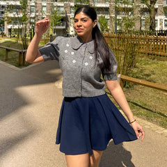 Woman in a gray cardigan with floral patterns and a navy blue skirt standing outdoors.