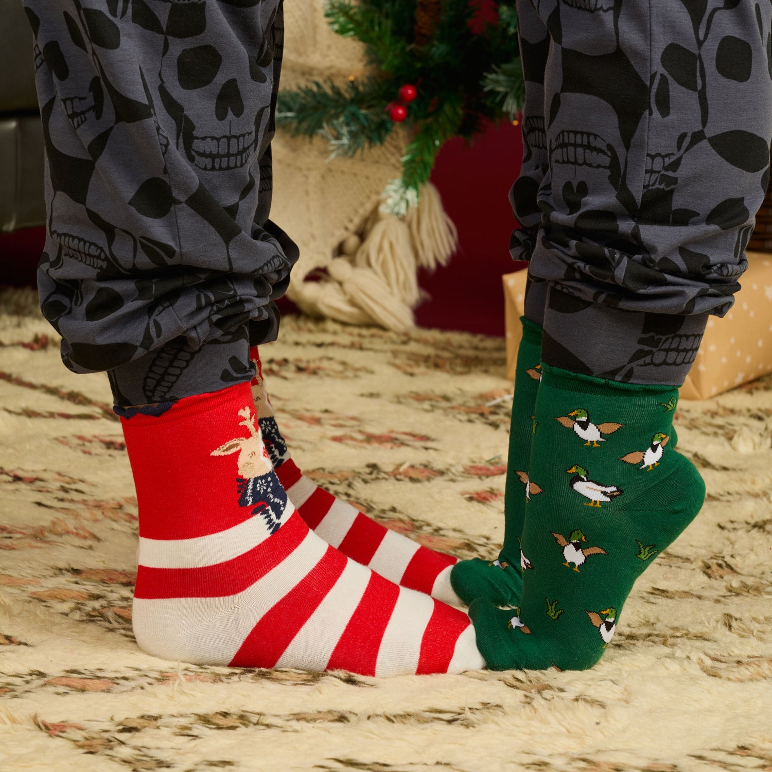 Close-up of feet on a rug wearing red striped Reindeer socks and green Duck bamboo socks, styled with grey skull pyjama cuffs.
